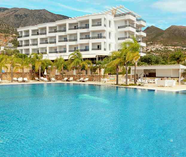 Et utendørsbasseng foran et hotell bygget i en moderne hvit design, omgitt av palmer og fjell i bakgrunnen. (AI-generert tekst)