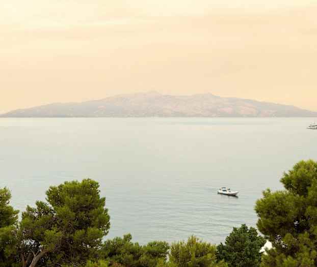 En fjern øy ses over havet med trær i forgrunnen og to båter på vannet. (AI-generert tekst)