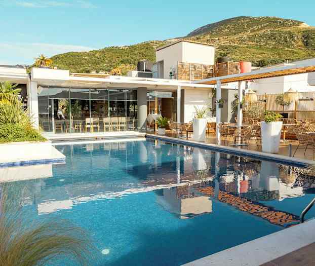 En moderne bygning med glassfasade ved siden av et utendørsbasseng, omgitt av grønn vegetasjon. (AI-generert tekst)