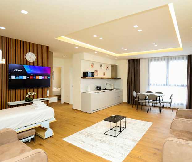 Et moderne hotellrom med åpen planløsning som inkluderer en sovesofa, TV, kjøkkenområde, spisebord og store vinduer med gardiner. (AI-generert tekst)