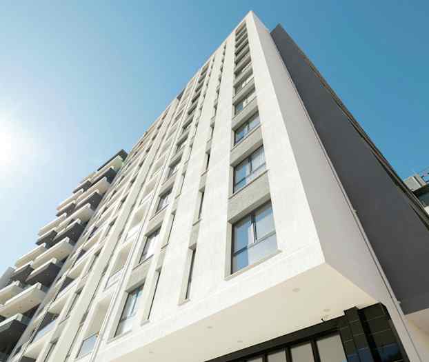 Bildet viser en moderne bygning med flere etasjer, fasaden er lys og har mange vinduer. En klar blå himmel og direkte sollys vises. (AI-generert tekst)