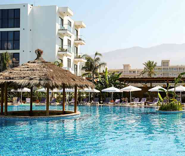 Et basseng med en stråtakspaviljong omgitt av solsenger og parasoller, med flerbyggingskomplekser i bakgrunnen. (AI-generert tekst)