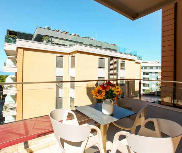 En balkong med utsikt over bygninger og havet. På balkongen er det et bord med en vase med blomster. (AI-generert tekst)