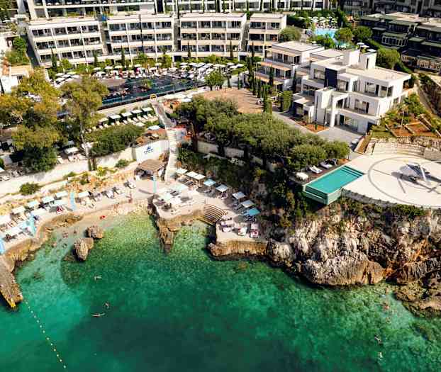 Luftfoto av et kysthotell med basseng, helikopterplass og strand mot turkis vann. (AI-generert tekst)