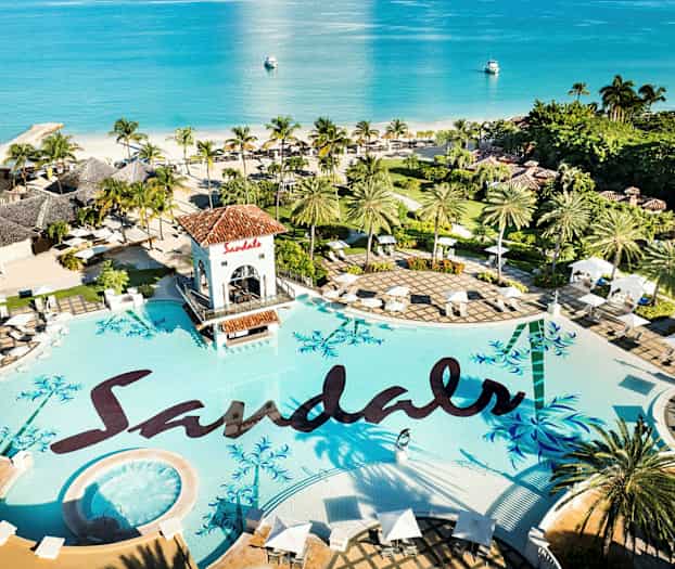 Et stort basseng med teksten 'Sandals', omgitt av palmer og parasoller, med utsikt over havet. (AI-generert tekst)