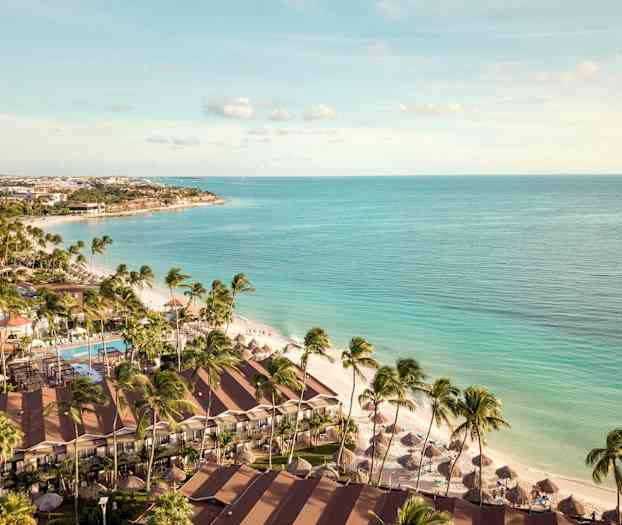 Luftfoto av en strand med palmer, hotellbygninger og parasoller nær klart blått hav. (AI-generert tekst)