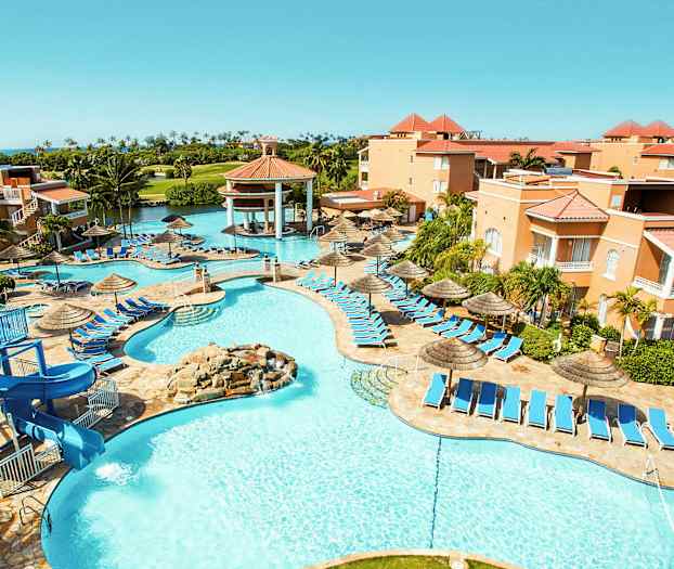 Bilde av et feriehotell med bassengområde, solsenger og parasoller. Bygninger omgir området og grønne palmer er synlige i bakgrunnen. (AI-generert tekst)