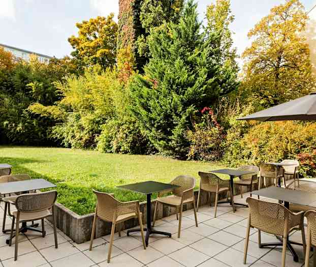 En utendørsrestaurant med bord og stoler på et fliselagt område ved siden av en grønn hage med busker og trær. (AI-generert tekst)