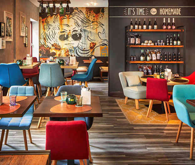 Et restaurantinteriør med fargerike sittemøbler, dekorative bord med drikker og bestikk samt en vegg med hyller fulle av forskjellige produkter. (AI-generert tekst)