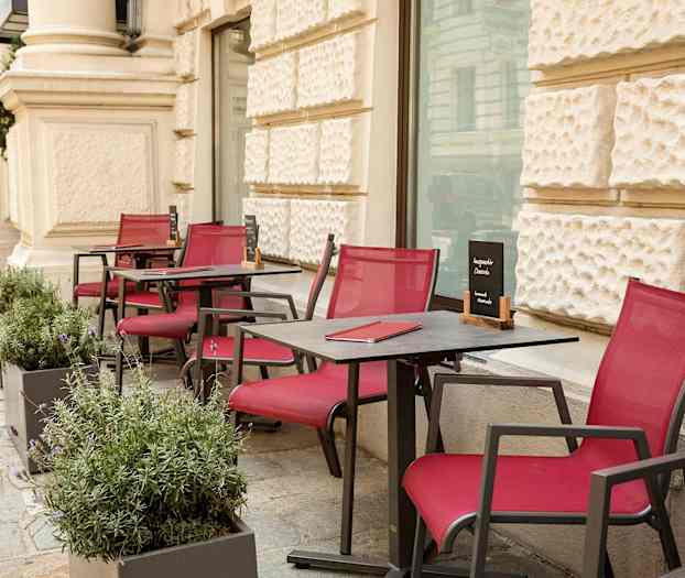 Utendørs bord og stoler med røde sitteputer, plassert langs en gangvei ved en bygning. (AI-generert tekst)