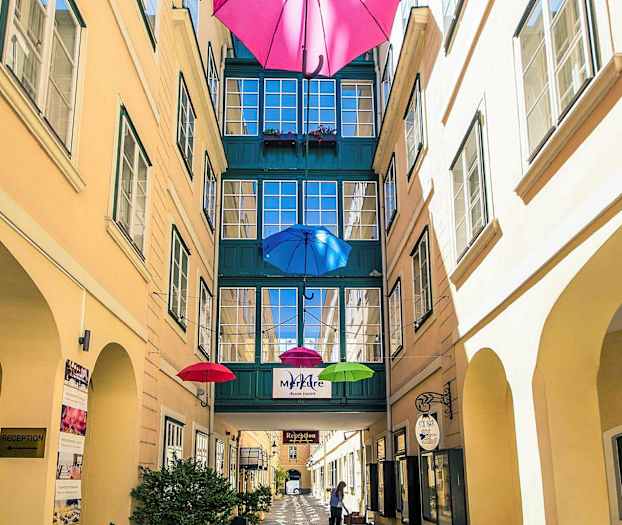 En smal gårdsplass med fargerike paraplyer hengende over og en passasje mellom bygninger. (AI-generert tekst)