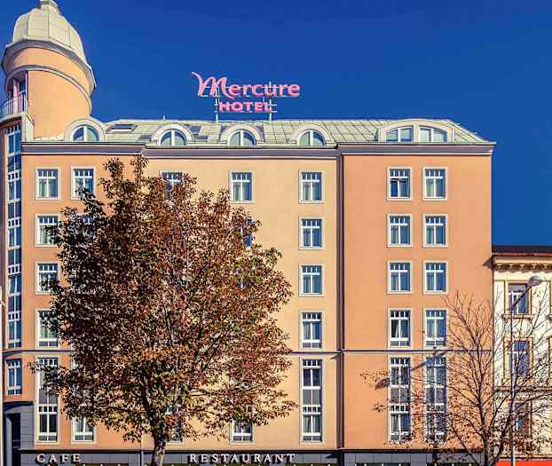 Eksteriøret av et hotell med navnet Mercure, med en beige fasade, mange vinduer og et høyt tårn på toppen. (AI-generert tekst)