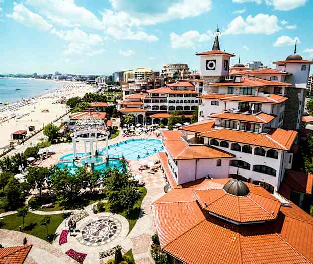 Et feriested med basseng, omgitt av bygninger med røde tak, ligger ved en strand med parasoller og solsenger. (AI-generert tekst)