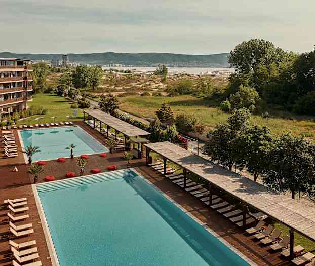 Stor basseng med solsenger og parasoller ved siden av et hotell, med grøntområder og landskap i bakgrunnen. (AI-generert tekst)