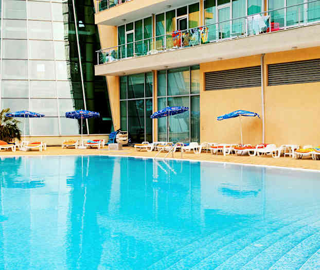 Et utendørsbasseng med solsenger og parasoller ved kanten, plassert foran en bygning med glassvinduer og balkonger. (AI-generert tekst)