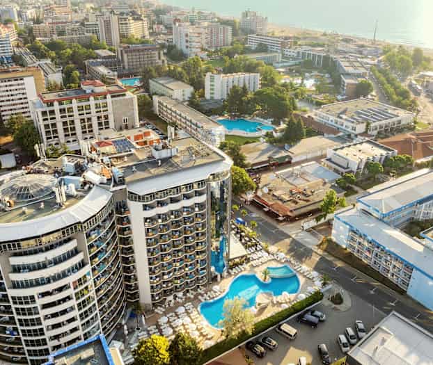 Luftfoto av hotellområde med basseng og omkringliggende bygninger nær sjøen. (AI-generert tekst)