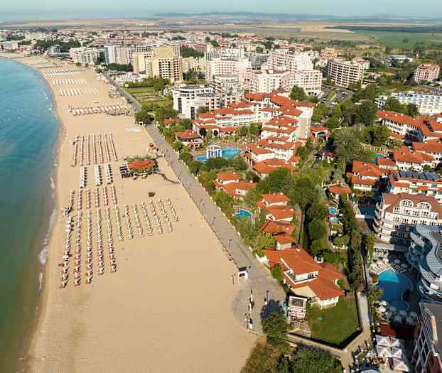 Utsikt over en strand med parasoller og solsenger samt omkringliggende bygninger. (AI-generert tekst)