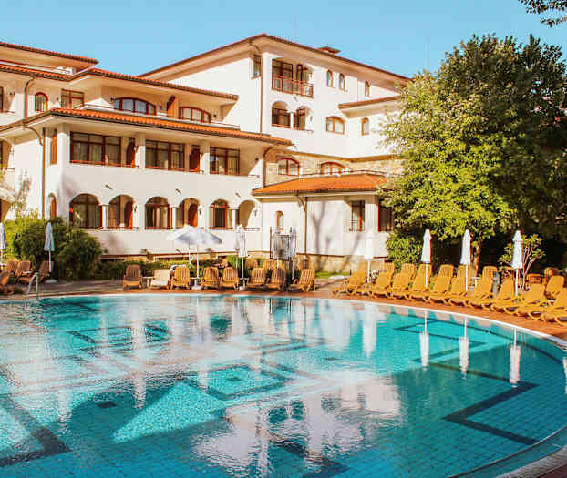 Et basseng foran en hvit bygning med røde tak, omgitt av solsenger og grønne trær. (AI-generert tekst)