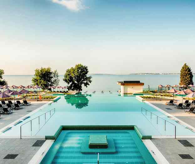 En utsikt over et basseng med solsenger og parasoller spredt rundt, samt havet og trær i bakgrunnen. (AI-generert tekst)