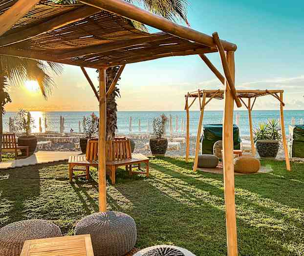 Utemøbler under treoverbygg nær stranden ved solnedgang. (AI-generert tekst)