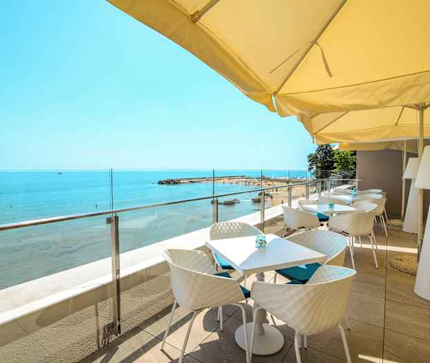 Terrasse med bord og stoler under parasoller som vender ut mot stranden og havet. (AI-generert tekst)