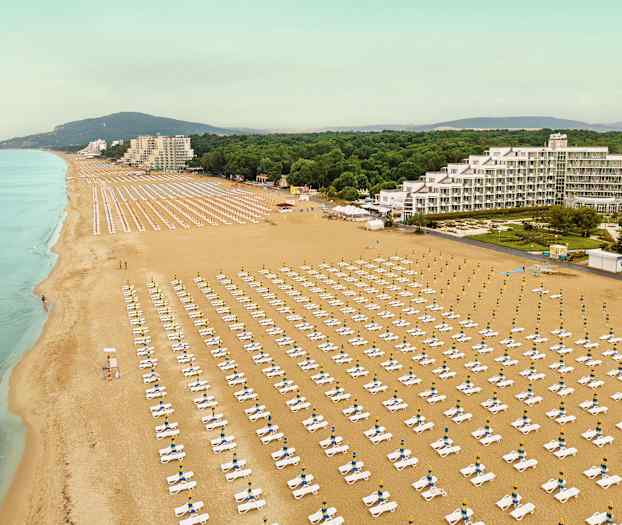 Flyfotografi av en stor sandstrand med rader av solsenger og parasoller, og bygninger og vegetasjon i bakgrunnen. (AI-generert tekst)