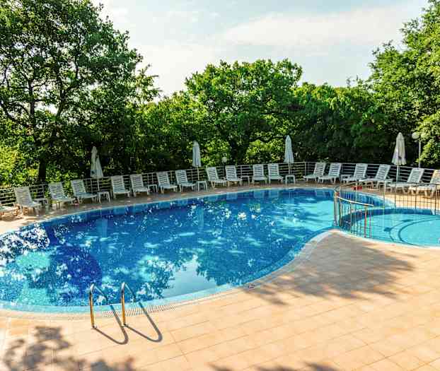 Bilde av et utendørs svømmebasseng omgitt av solsenger og parasoller, med grønne trær i bakgrunnen. (AI-generert tekst)