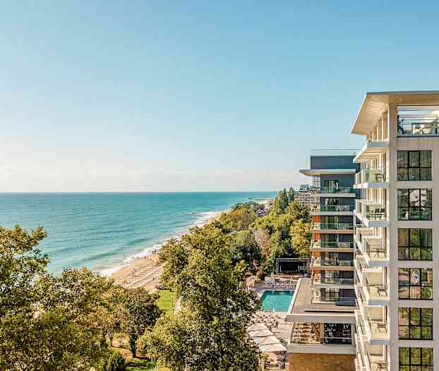 En utsikt fra høyden over kysten med et hotell til høyre og stranden omgitt av trær. (AI-generert tekst)