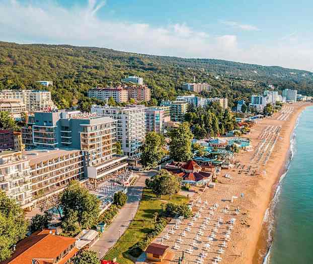 Bilde av en kystby med sandstrand, parasoller og bygninger ved grønne åser. (AI-generert tekst)