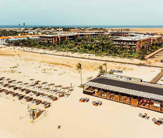 Flyfoto av en sandstrand med parasoller og solsenger, samt en strandrestaurant med bord og stoler, omgitt av resortbygninger og grønne planter. (AI-generert tekst)