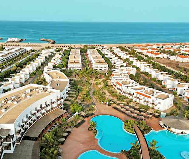 Luftfoto av et hotellområde med bassenger, bygninger og strand i bakgrunnen. (AI-generert tekst)