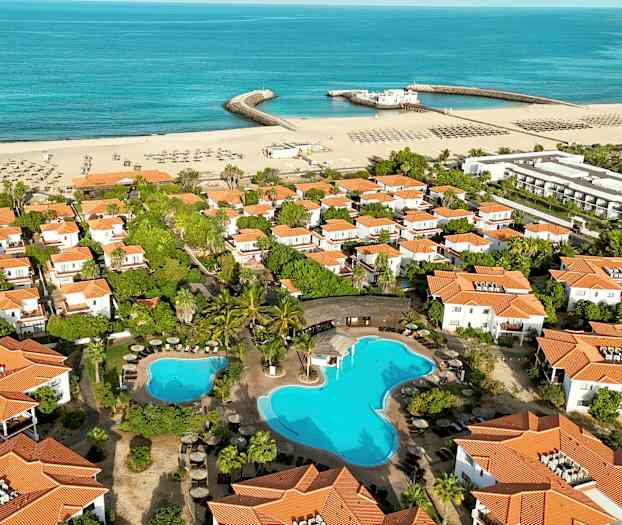 Flyfoto av et ferieområde med hvite hus med oransje tak, svømmebasseng og en sandstrand ved havet. (AI-generert tekst)