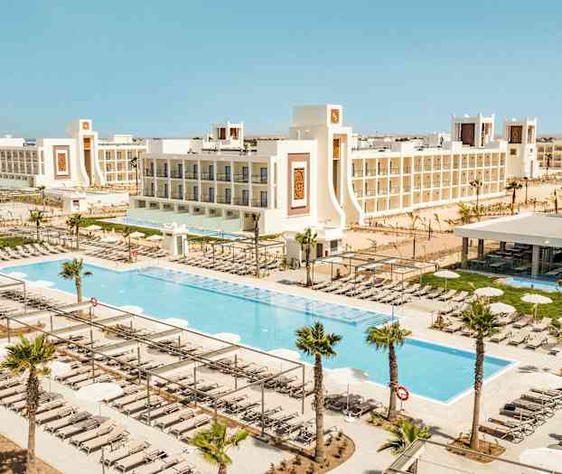 En utsikt over et feriested med bassenger, solsenger og omgitt av hvite bygninger. (AI-generert tekst)