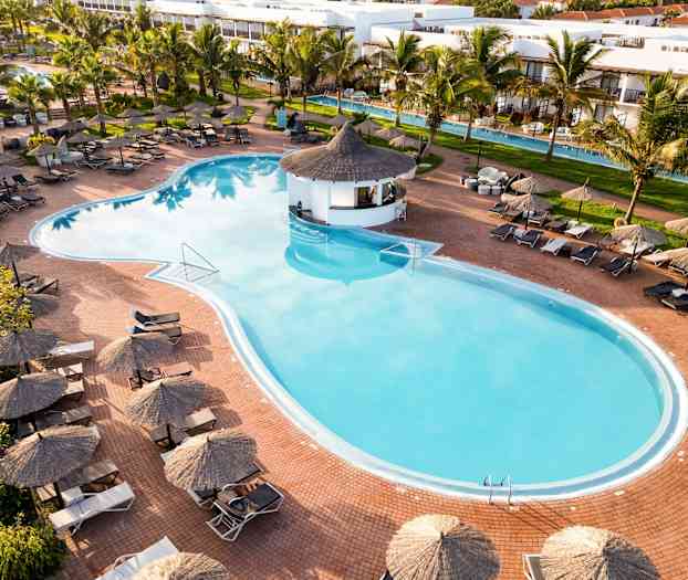 En utsikt over et stort utendørsbasseng med solsenger og parasoller rundt, omgitt av palmer og en frodig hage. (AI-generert tekst)