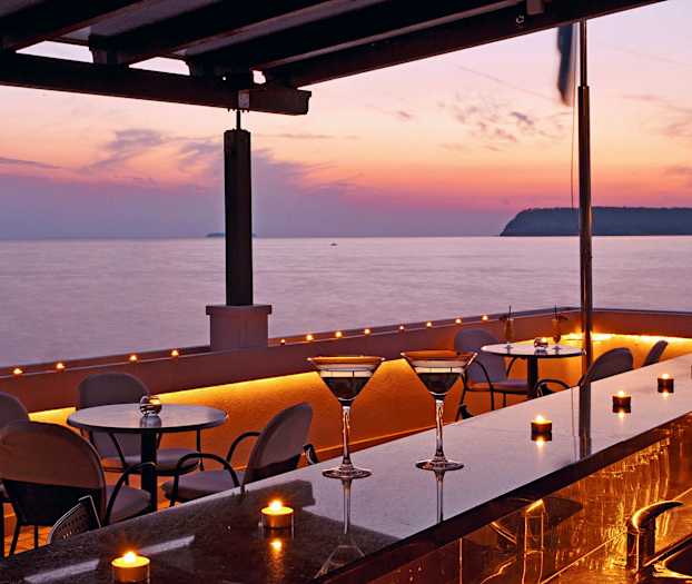 Uteservering med bord og stoler ved sjøen i solnedgangen. (AI-generert tekst)