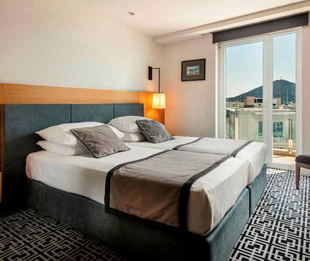 Hotellrom med dobbeltseng, lys innredning og balkong med utsikt over by og fjell. (AI-generert tekst)