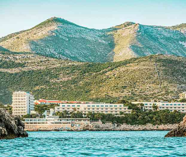 En kystutsikt med hav i forgrunnen, bygninger i midten og grønne fjell i bakgrunnen. (AI-generert tekst)