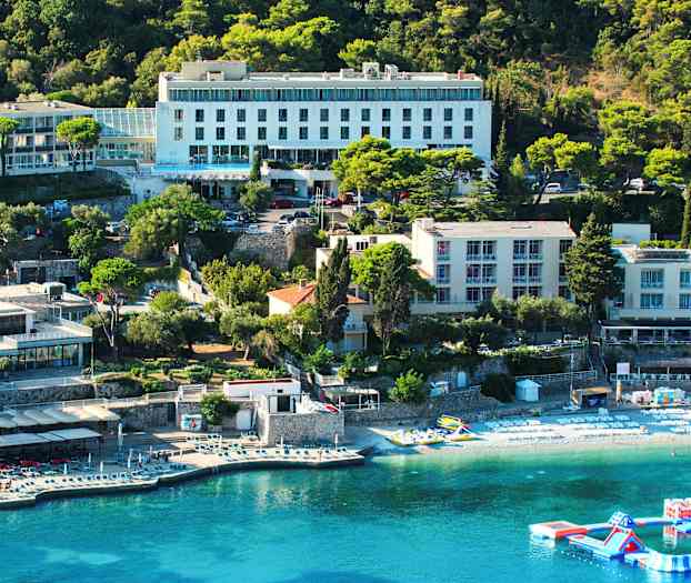 En utsikt over et hotellbygning ved en kystlinje med solsenger på stranden og en flytende lekeplass i vannet. (AI-generert tekst)