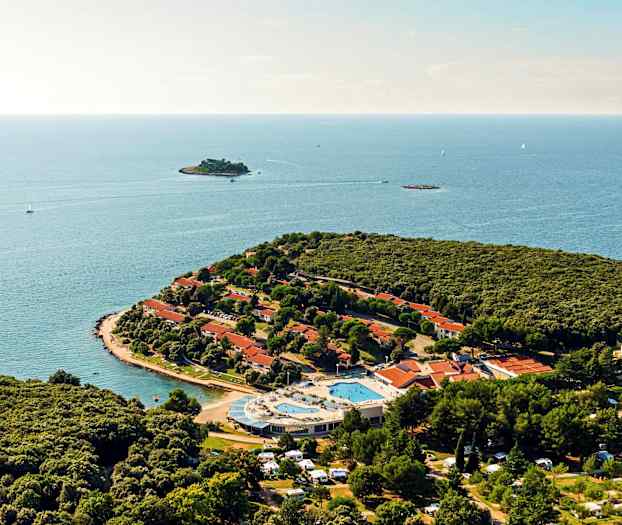 Flyfoto av en halvøy med tett vegetasjon, røde takhus, et bassengområde og det omkringliggende havet. (AI-generert tekst)