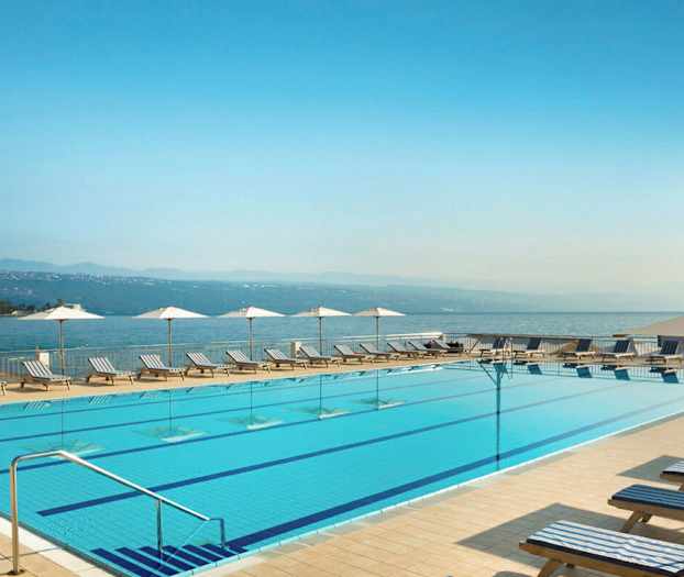 Et utendørsbasseng med solsenger og parasoller langs kanten. I bakgrunnen er havet og fjellene synlige. (AI-generert tekst)