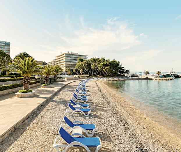 En steinstrand med solsenger og palmer, omgitt av hotellbygninger og trær. (AI-generert tekst)