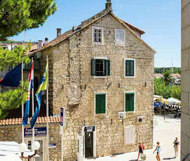 En steinbygning med grønne vinduslemmer og flagg foran, omgitt av trær og mennesker som spaserer. (AI-generert tekst)