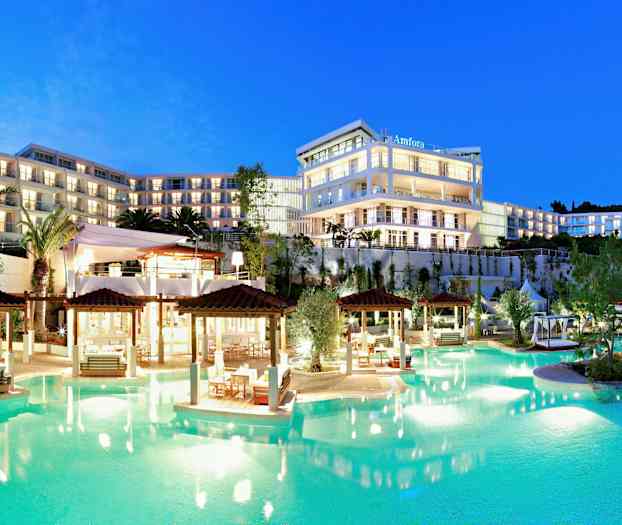 Et opplyst hotellkompleks med flere etasjer ved et basseng, omgitt av hager og palmer, i skumringen. (AI-generert tekst)