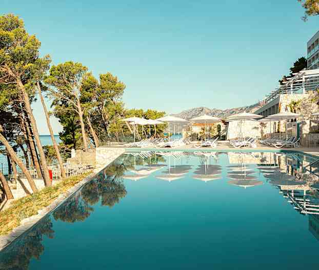 Utsikt over et infinitybasseng med solsenger og parasoller langs kanten, omgitt av trær og bygninger. (AI-generert tekst)