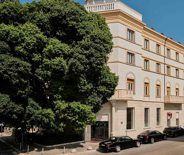 En bygning med flere etasjer og klassisk stil med buede vinduer og en fasade i lys farge. Foran bygningen står biler parkert ved siden av et stort frodig tre. (AI-generert tekst)