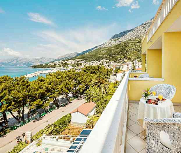 Utsikt fra en balkong på en gul bygning over en by ved havet og omkringliggende fjell. (AI-generert tekst)