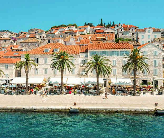 Bilde av en by ved vannet med teglrøde tak og palmer langs strandpromenaden. (AI-generert tekst)