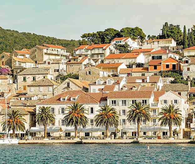 Bildet viser en pittoresk by ved sjøen med røde teglsteinstak, steinhus og palmer langs kaien. (AI-generert tekst)