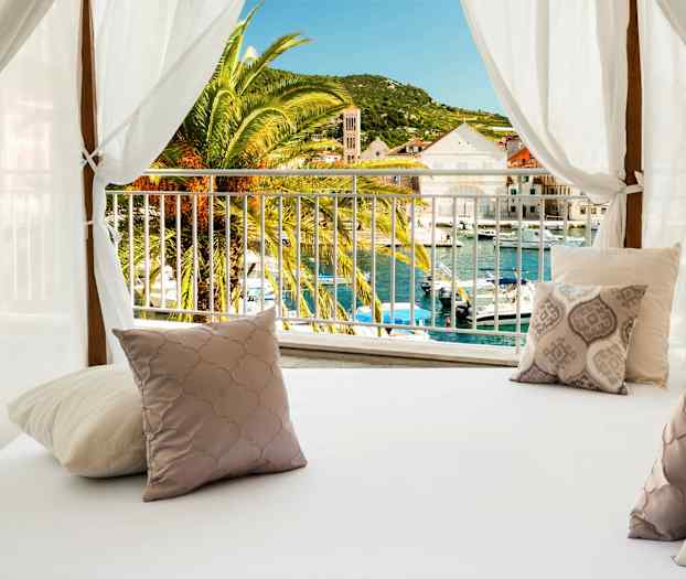 En seng med dekorative puter under en hvit baldakin med utsikt over en palme og en havn. (AI-generert tekst)