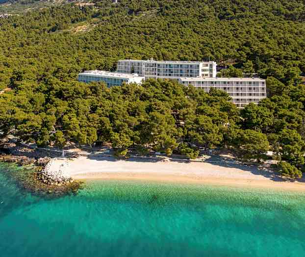 Hotellbygninger omgitt av grøntområder nær en strand med klart turkist vann. (AI-generert tekst)